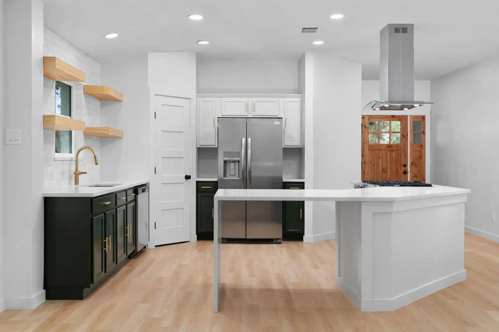 South-facing Austin kitchen remodel with deep green base cabinets, brass hardware, floating oak shelves and a quartz-topped island &mdash; Bremner full remodel by Bailey & Bear Construction