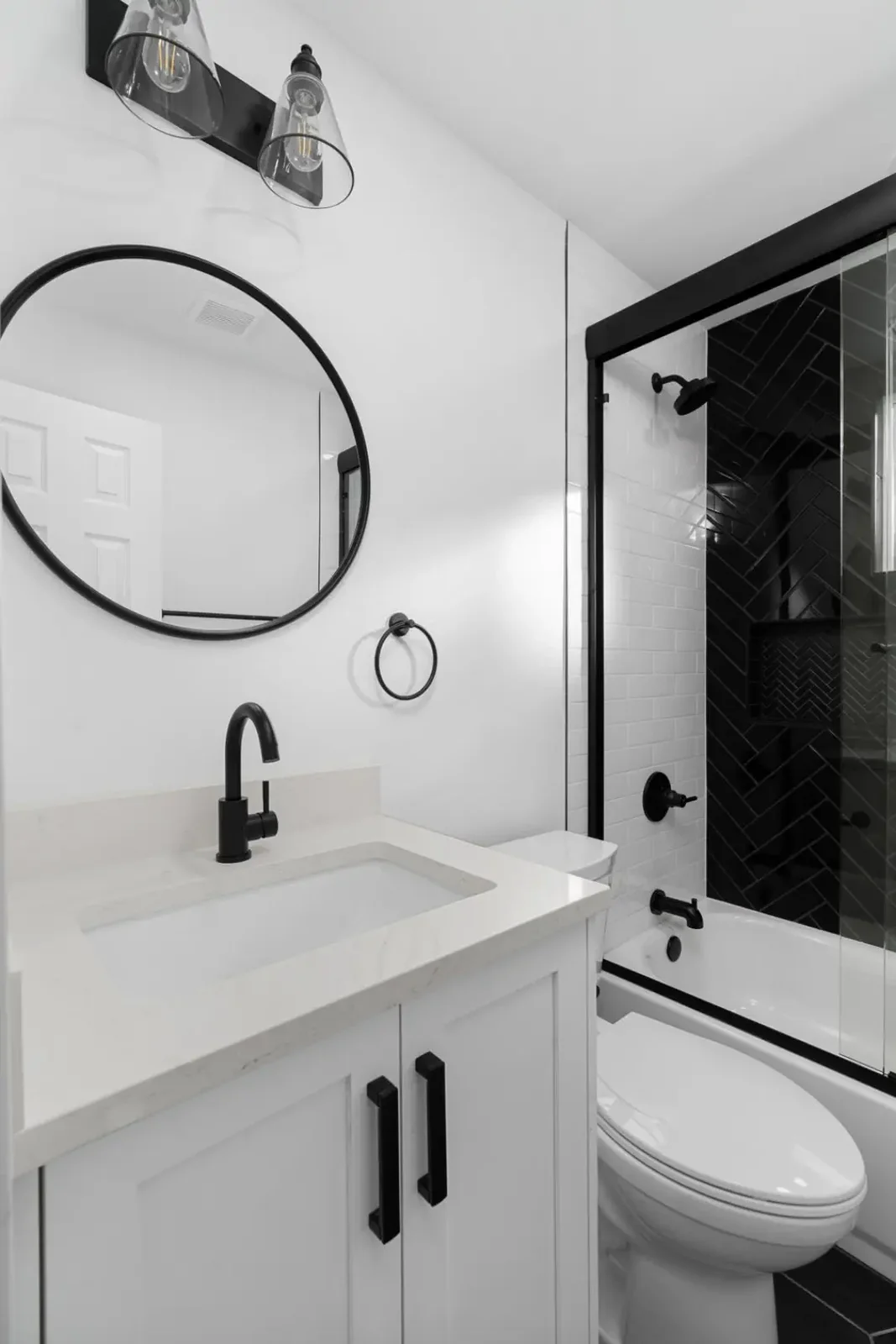 Compact remodeled guest bathroom with a white quartz-topped vanity, round black mirror, matte black fittings and a tiled tub-shower &mdash; Emerald Forest remodel in Austin by Bailey & Bear Construction